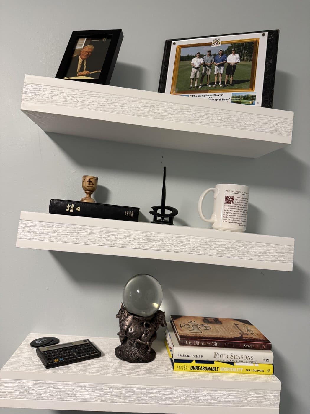 Three white shelves in the Bob Bingham room: a framed photo of Bob at his desk and a golf photo of the Bingham men on top, a Bible and a Prudent Man Rule mug in the middle, and on the bottom his HP 12c calculator, a crystal ball on a bronze bull and bear stand, and books including Isadore Sharp's Four Seasons.