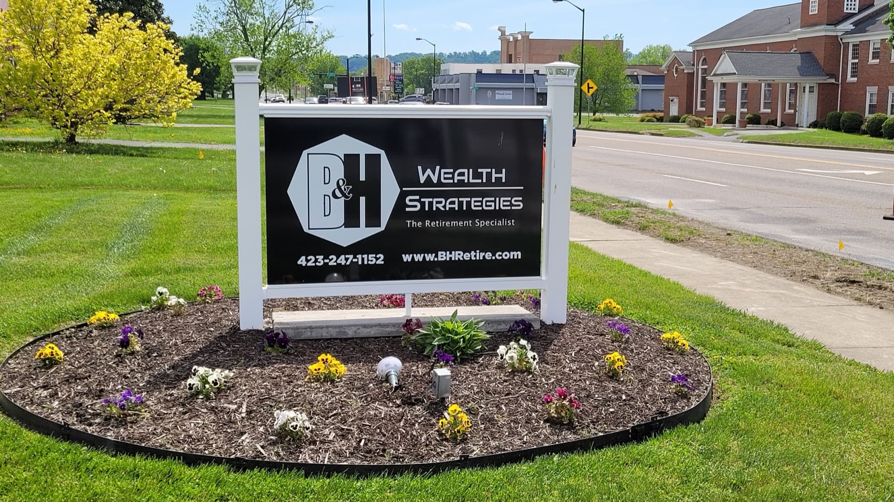 The B&H Wealth Strategies sign in its flower bed on Center Street, with the road and traffic behind it.