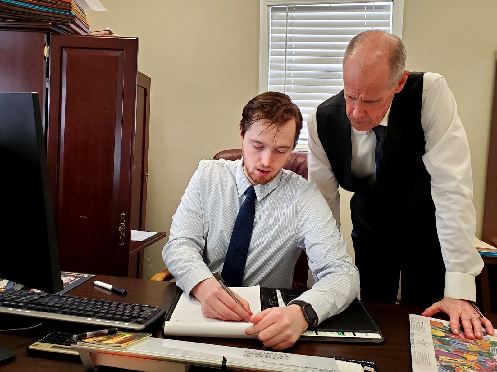 Jeff Bingham leaning over the desk beside Jake, who is writing on a legal pad, the two of them working through something together in the Kingsport office.