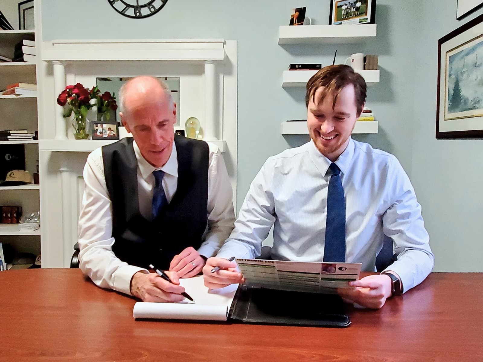 Jeff and Jake Bingham at the office table, Jeff writing on a legal pad while Jake looks over a page of figures, both of them smiling.